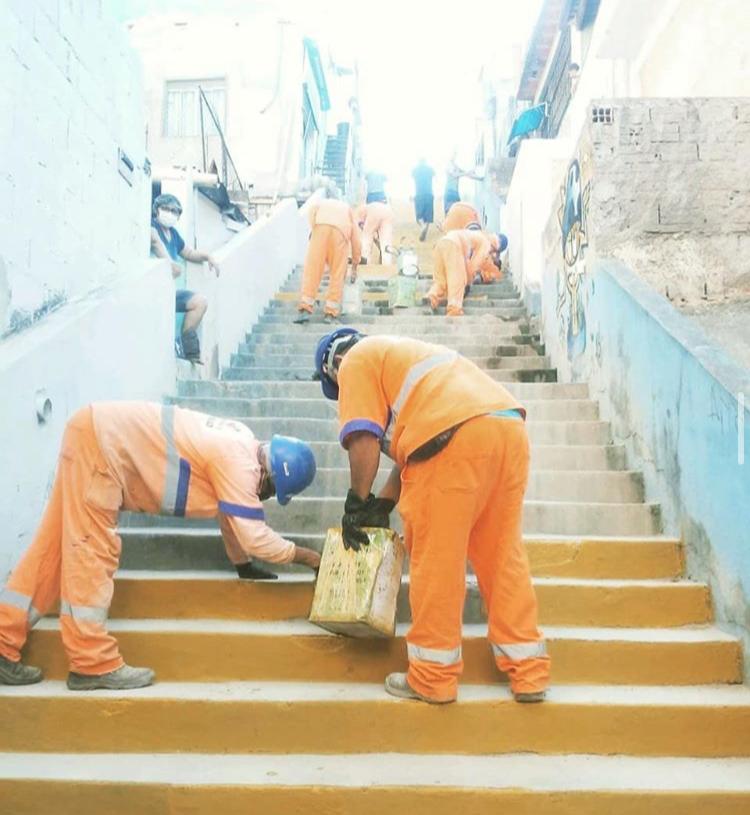#PraTodosVerem visualiza-se trabalhadores, vestidos de uniformes da cor laranja, pintando uma escada de amarelo.